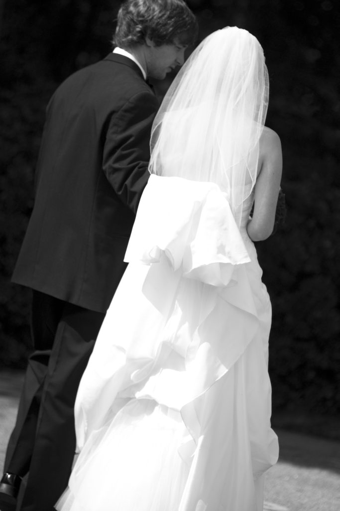 A photo from behind of a newlywed couple walking. The man on the left in a black tuxedo with his arm around the back of a forty year old woman dressed in a white wedding dress and veil.