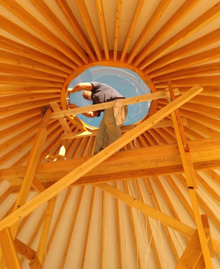 Man standing on scaffolding in the center of a yurt installing the dome