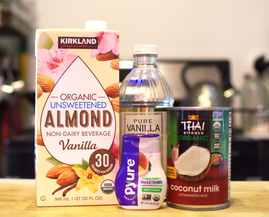 A box of almond milk, can of coconut milk, a bottle of vanilla, and a squirt bottle of organic sweetener on a counter